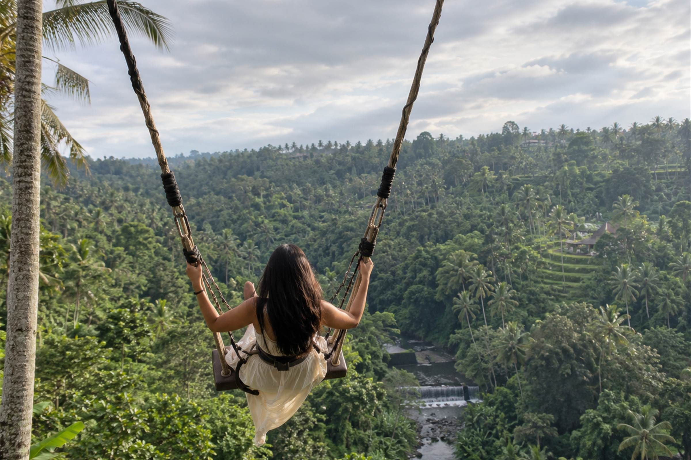A traveller on a rope swing above a palm-filled valley and rice terraces near Ubud